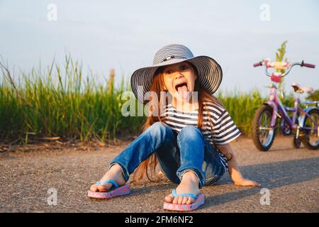 Carino bambina che mostra la lingua fuori nel cappello grande del sole seduto su strada con la sua bicicletta dietro di lei Foto Stock