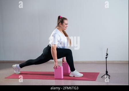 Giovane donna caucasica grassa affondo su un tappetino sportivo. Affascinante più la ragazza di taglia in sportswear sta facendo le esercitazioni di idoneità con un addestratore in linea sul telefono Foto Stock