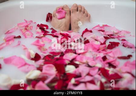 Divertente immagine di un uomo che prende un bagno rilassante. Primo piano dei piedi maschi in un bagno con schiuma e petali di rosa Foto Stock