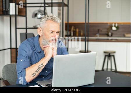 Uomo di mezza età serio in camicia casual intelligente perso nei pensieri seduti alla scrivania con un computer portatile e lavorando a distanza da casa, capelli grigi maschio riposa mento con entrambe le mani e fissando allo schermo Foto Stock