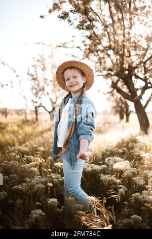 Ragazza di 4-5 anni di stile che indossa cappello di paglia e abiti alla moda in denim in prato all'aperto. Guardando la fotocamera. Infanzia. Stagione primaverile. Foto Stock