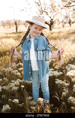 Felice ragazza di 4-5 anni con cappello di paglia e giacca decnim con pantaloni in posa in fiore prato all'aperto. Guardando la fotocamera. Stagione primaverile. Childho Foto Stock