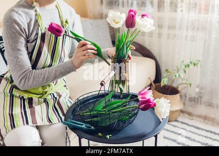 Donna mette bouquet di fiori tulipani in vaso con acqua a casa. Fiori freschi bianchi e viola raccolti nel cestino. Interni e decorazioni Foto Stock