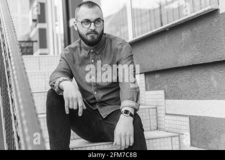 Immagine in scala di grigi di un maschio biondo caucasico con barba e bicchieri che posano seduti sulle scale Foto Stock