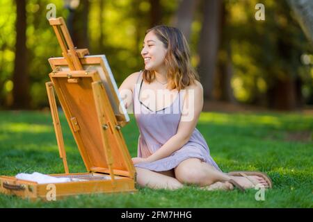 Petite giovane donna asiatica studente Pittura nel Parco utilizzando Un armadietto Easel Foto Stock