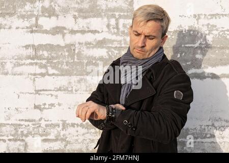 Affascinante uomo biondo di mezza età di aspetto modello di fronte a una parete di mattoni bianchi. L'uomo guarda il tempo sul suo orologio da polso. Modello maschio Foto Stock