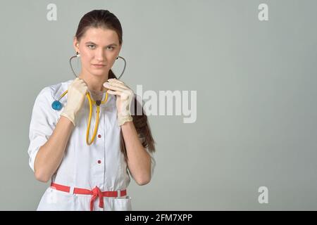 Una giovane dottoressa in un camice bianco da laboratorio con uno stetoscopio in mano su uno sfondo grigio, un posto per il testo Foto Stock