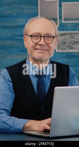 Pensionato manager seduto alla scrivania di fronte alla macchina fotografica sorridente dopo aver digitato al computer portatile lavorando da casa, mentre la moglie anziana leggere un libro in background. Uomo preparato per meeting online utilizzando gadget moden Foto Stock