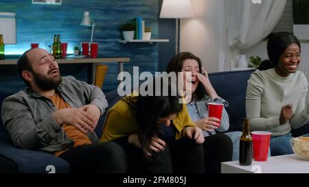 Amici multietnici che hanno una reazione divertente mentre si guarda film comici godendo di una serata di film. Gruppo misto di persone che mangiano spuntini, bere birra divertendosi insieme durante la festa serale Foto Stock