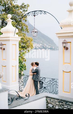 Gli sposi novelli abbracciano e quasi baciano sotto un vecchio arco sullo sfondo del Lago di Como. Vista laterale Foto Stock