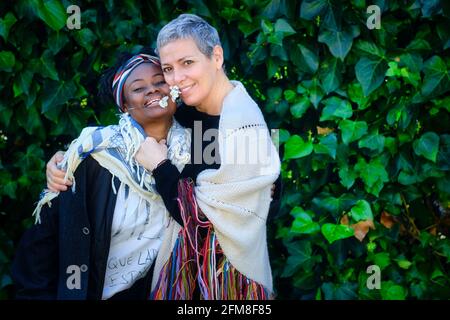 Donna caucasica matura e giovane donna africana che abbracciano in un giardino con un background di foglie. Foto Stock