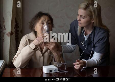 Giovane medico di donna molto abile che mette sulla maschera nebulizzatrice sopra faccia della sua paziente femminile anziana per fare l'inalazione quando visitarlo a casa Foto Stock