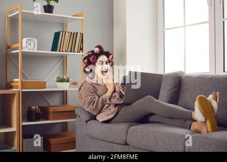 Sorpresa eccitato giovane donna seduta sul divano e guardando il suo computer portatile Foto Stock