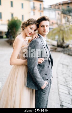 Sposa abbraccia lo sposo da dietro, appoggiando la testa sulle spalle, in piedi sul lungomare del Lago di Como Foto Stock