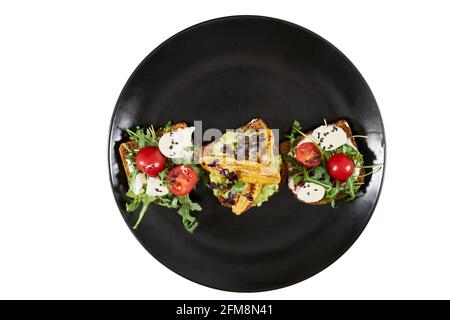 Vista dall'alto del bellissimo piatto nero moderno con appetitosi toast con pomodori, mozzarella e rucola su sfondo bianco. Concetto di spuntini sani e gustosi a casa. Foto Stock