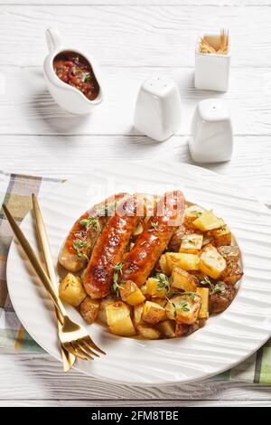 bangers e patate arrostite su un piatto bianco con sugo di cipolla in una caraffa su un tavolo di legno testurizzato bianco, vista verticale dall'alto Foto Stock