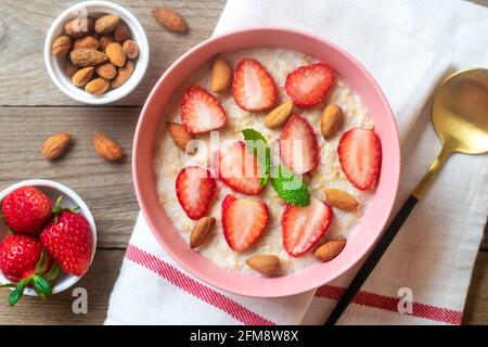 Porridge di farina d'avena con fragole, mandorle, foglie di menta in ciotola rosa, cucchiaio e tovagliolo con strisce rosse su sfondo di legno Vista dall'alto Flat Lay Vegan Foto Stock