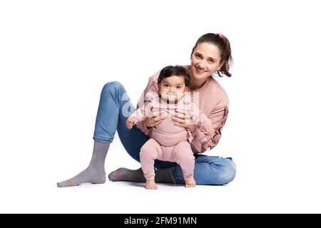 Ritratto di madre carina con figlia che guarda la fotocamera isolata su sfondo bianco studio. Giovane donna attraente seduta sul pavimento e aiutando dolce adorabile bambino a stare in piedi, felice concetto di infanzia. Foto Stock