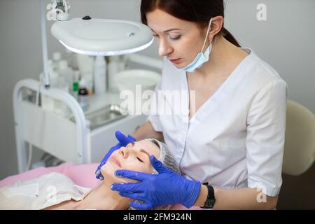 Primo piano della giovane confidente dottoressa cosmetologa, vagando uniforme bianca e guanti sterili blu, applicando schiuma detergente sul viso della giovane donna, sdraiata sul divano. Cura della pelle Foto Stock