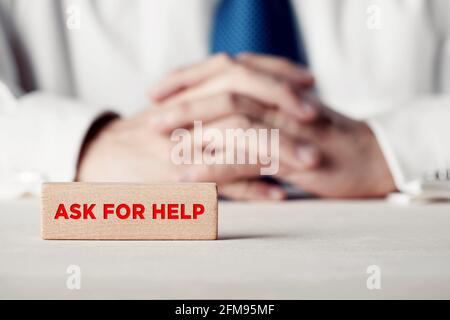 Chiedi un messaggio di aiuto scritto su un blocco di legno di fronte a un uomo d'affari con mani incrociate. Assistenza, consigli, indicazioni per clienti o professionisti Foto Stock