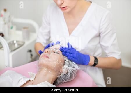 Primo piano di medico dermatologo professionale in guanti di gomma blu, applicando pellicola sul viso di giovane paziente donna per ringiovanimento della pelle e idratazione. Concetto di trattamenti per il viso Foto Stock