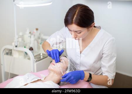 Primo piano di medico dermatologo professionale in guanti di gomma blu, applicando pellicola sul viso di giovane paziente donna per ringiovanimento della pelle e idratazione. Concetto di trattamenti per il viso Foto Stock