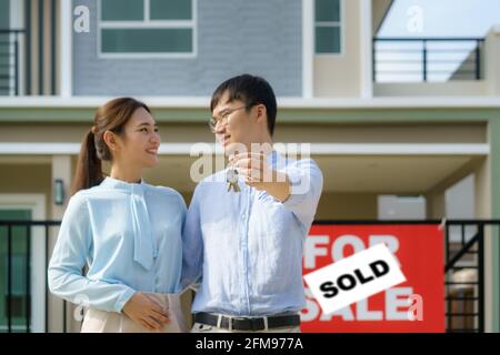 Ritratto di giovane coppia asiatica in piedi e abbracciando insieme e tenendo la chiave della casa che guarda felice davanti alla loro nuova casa per iniziare la nuova vita. Famiglia Foto Stock