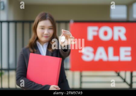 Agente immobiliare asiatico o donna immobiliare sorridente e tenendo file rosso con mostrare la chiave di casa di fronte alla casa. Foto Stock