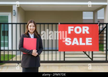 L'agente immobiliare delle donne asiatiche è in piedi di fronte la casa con segno di vendita appeso alla porta di casa a. annunciare che le parti interessate sono in co Foto Stock