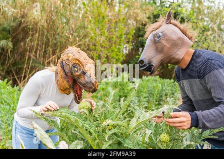 Felice coppia uomo e donna con dinosauro e testa di cavallo Maschera in verdure giardino. Fantasia e stile di vita umorismo maschile e. donna cercando di mangiare cibo outd Foto Stock