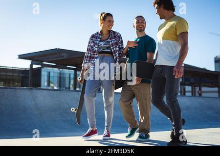 Tre amici caucasici felici che camminano con skateboard e parlando al sole Foto Stock