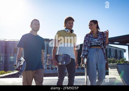 Tre amici caucasici femminili e maschili che camminano con skateboard e. parlare al sole Foto Stock