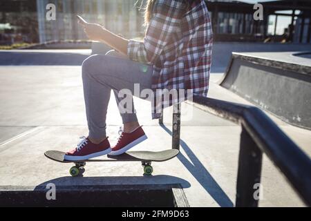 Sezione bassa della donna caucasica seduta sul corrimano con skateboard al sole Foto Stock