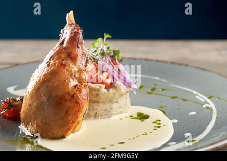 Primo piano di succosa gamba di pollo grigliata servita con purè di patate e funghi, germogli verdi freschi, pomodori ciliegini, panna acida e salsa verde. Bella piastra blu in ceramica su tavolo di legno. Foto Stock