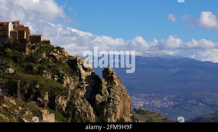 Italia, Sicilia, vista di una parte del villaggio Cesaro, situato su una collina, promontorio roccioso, nel centro sottostante è la città di Randazzo. L'Etna innevata sorge sullo sfondo, il cielo è azzurro con nuvole bianco-grigie Foto Stock