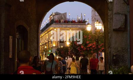 Italia, Sicilia, Taormina, centro città, umore serale, edificio storico illuminato, vista attraverso l'arco, passeggiate per gruppi di persone, terrazza sul tetto sullo sfondo Foto Stock