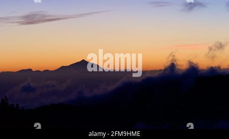 Spagna, Isole Canarie, Gran Canaria, Massiccio Centrale, montagne all'interno dell'isola, tramonto, cielo blu sopra, orizzonte rosso, Teide riconoscibile come una silhouette, nuvole scure in primo piano Foto Stock