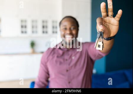 Fuoco selettivo sui tasti con portachiavi in forma di piccola casa in mano maschile. L'uomo afroamericano sorridente tiene le chiavi dalla nuova proprietà, compratore felice della propria proprietà. Un ragazzo si è rilocato Foto Stock