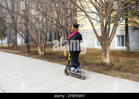 Giovane donna da dietro in abito nero graduato e nero berretto graduato con nappina gialla che guida uno scooter elettrico dopo laurea all'univer Foto Stock