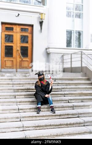 Studentessa giovane donna in abito nero di laurea, tappo di laurea con nappina gialla, maschera protettiva seduta su gradini di istituto educativo e. Foto Stock