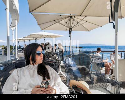 Vista frontale di una giovane donna seduta e con il telefono cellulare sulla terrazza in un caffè sul mare a Opatija. Foto Stock