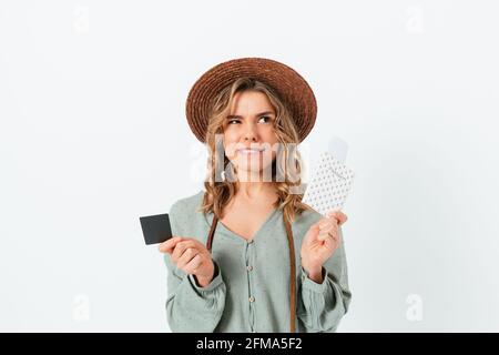 Ritratto di giovane donna in dubbio che indossa il cappello estivo che tiene il passaporto con i biglietti di viaggio e la carta di credito, guardando in alto, in piedi sulla schiena bianca Foto Stock