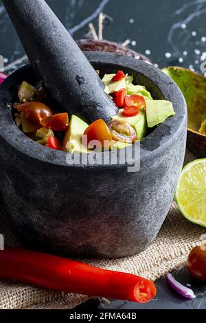 Preparazione di guacamole in un tradizionale mortaio di pietra con tutti i suoi ingredienti (avocado tritato, lime, cipolla, pomodori e peperoncini). Appetizin Foto Stock