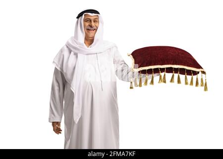 Sorridente arabo maturo in un accappatoio bianco e un copricapo tenendo un cuscino di velluto rosso isolato su sfondo bianco Foto Stock