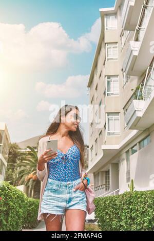 Giovane donna bionda in tuta rossa e con maschera protettiva per il viso scattando una foto selfie vicino al viale di lusso e alla spiaggia. Nuovo concetto normale Foto Stock