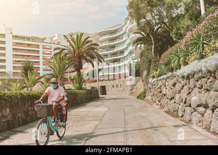 Padre con maschera in bicicletta con suo figlio in un seggiolino, mattina a piedi sul viale, giorno di sole. Nuovo concetto normale Foto Stock