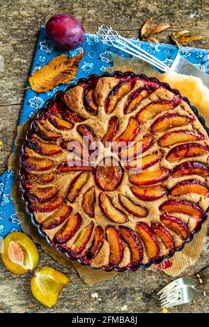 Torta di prugne su sfondo rustico di legno, panno fiorito blu, prugne tagliate, foglie d'autunno, una prugna fresca, posate. Foto Stock