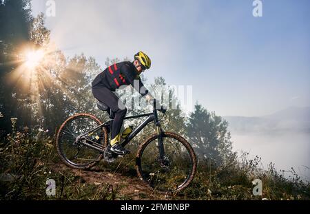 Immagine orizzontale dell'uomo che guida la sua bicicletta in montagna in mattinata in salita. Il sole splende attraverso la corona di alberi, si innevate sulle nuvole sullo sfondo. Concetto di stile di vita attivo Foto Stock