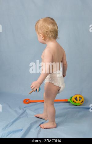 Un piccolo ragazzo biondo di 1 anno in un pannolino bianco si erge su uno sfondo blu. Vista dal retro Foto Stock