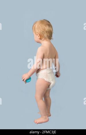 Una bambina bionda di 1 anno in un pannolino bianco si erge su uno sfondo blu. Vista dal retro Foto Stock